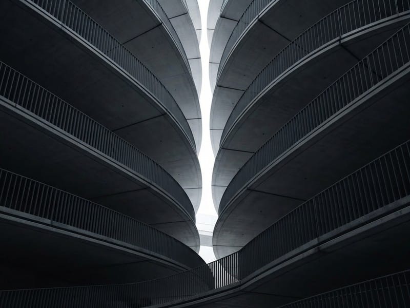 Curving concrete parking ramps with metal railings rise upward on both sides, meeting near a vertical opening of daylight.